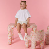 Young girl sitting on a floral-patterned stool against a pink background