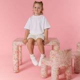 Young girl sitting on a floral-patterned stool against a pink background