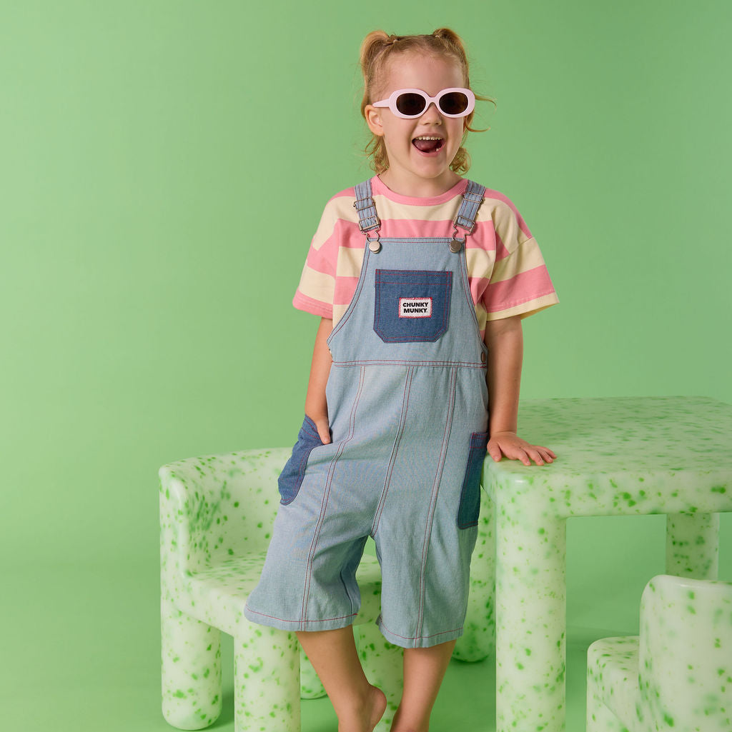 Child wearing sunglasses and a striped shirt with overalls standing on a green background