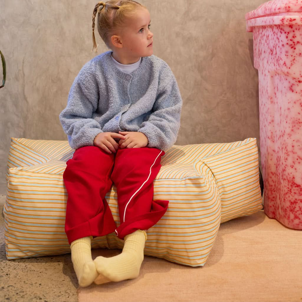Child sitting on a striped cushion against a textured wall with a pink object in the corner.