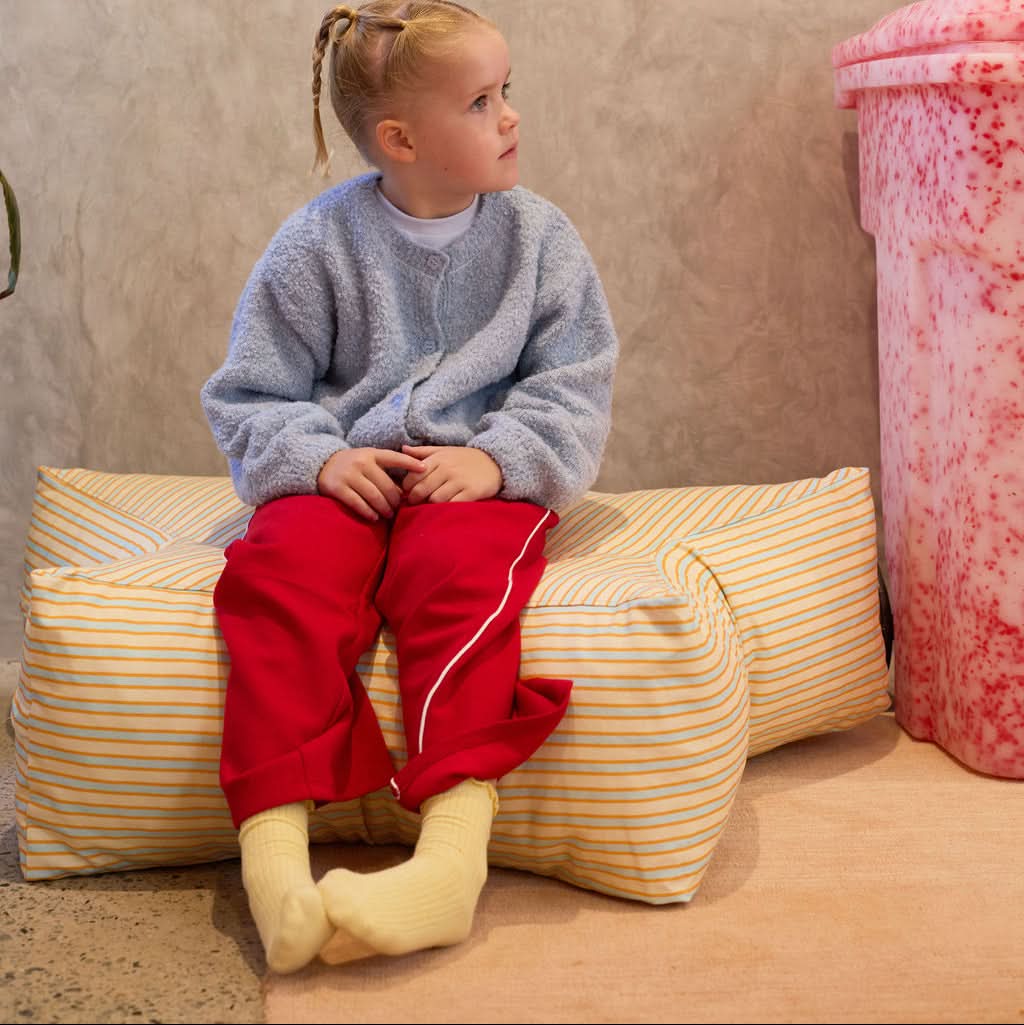 Child sitting on a striped cushion against a textured wall with a pink object in the corner.