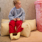 Child sitting on a striped cushion against a textured wall with a pink object in the corner.