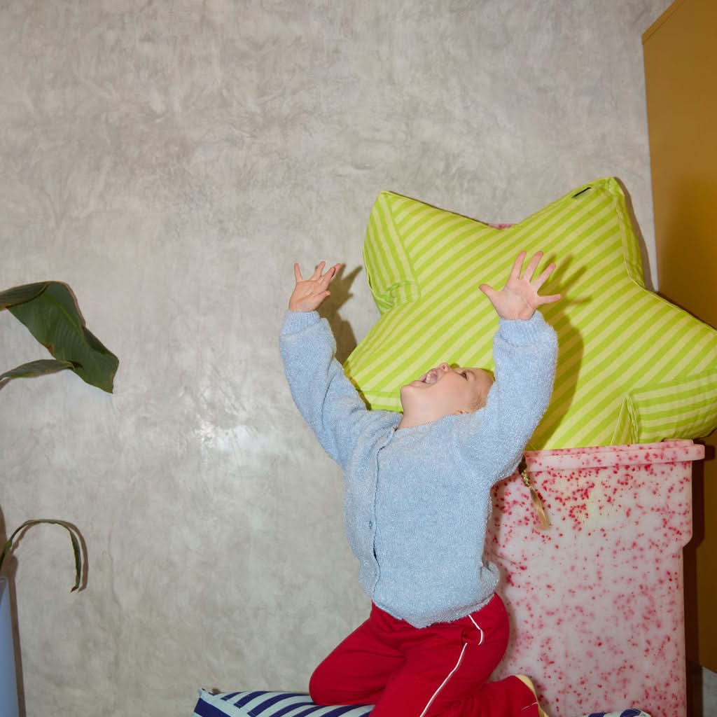 Child playing with colorful cushions on a striped ottoman against a textured wall.