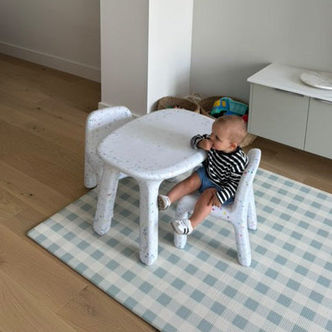 Child sitting on a small white chair in a room with wooden flooring and a checkered rug.