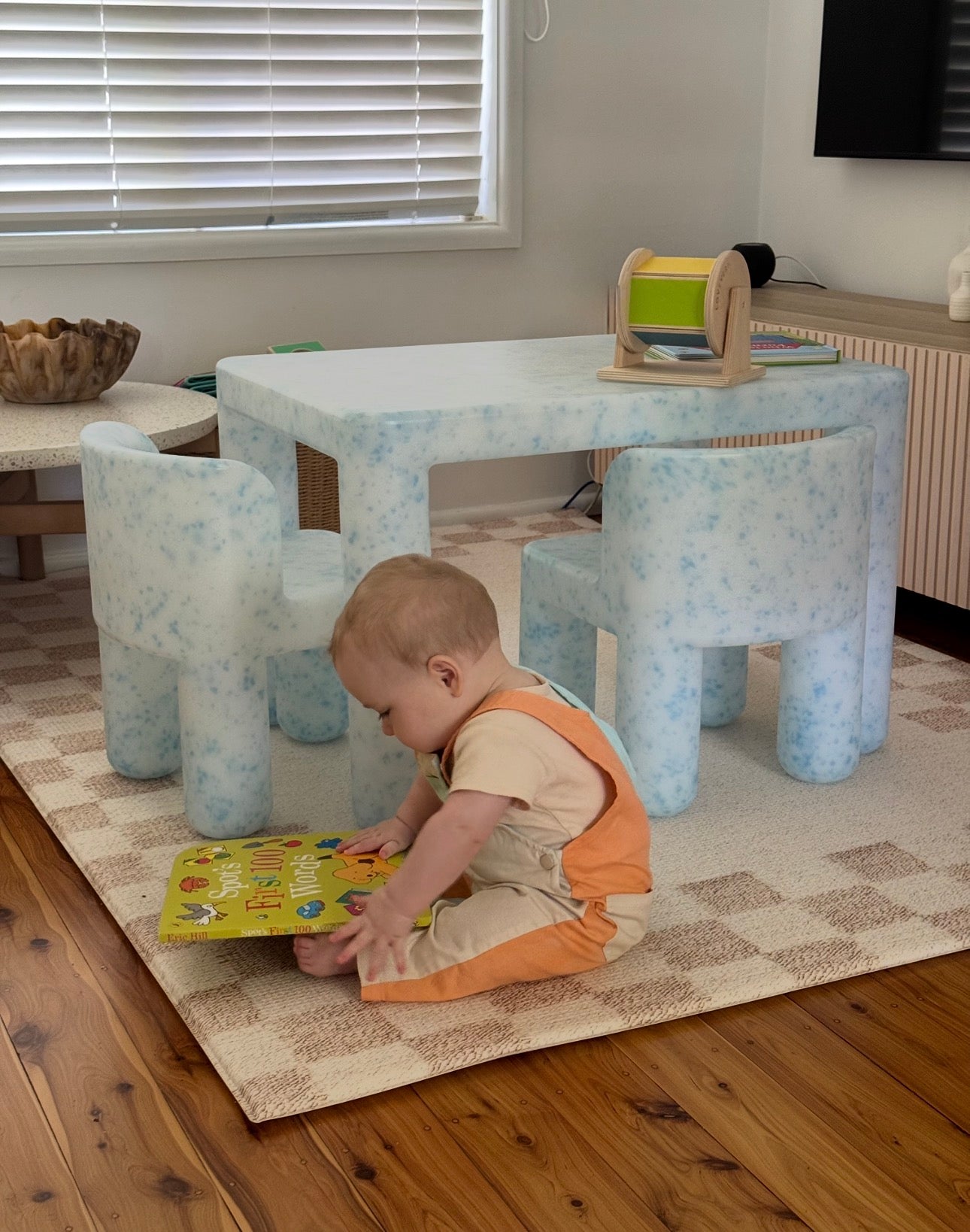 Child playing with a book on the floor near a children's table and chairs.
