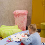 Child sitting on a striped ottoman at a small table with art supplies, in a room with colorful furniture.