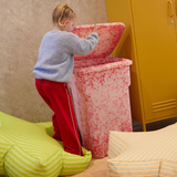 Child interacting with a large pink trash bin in a room with a yellow cabinet and green and yellow striped furniture.