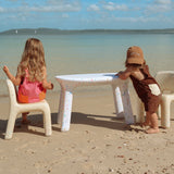 Two children sitting at a small table on a sandy beach with ocean and sky in the background.