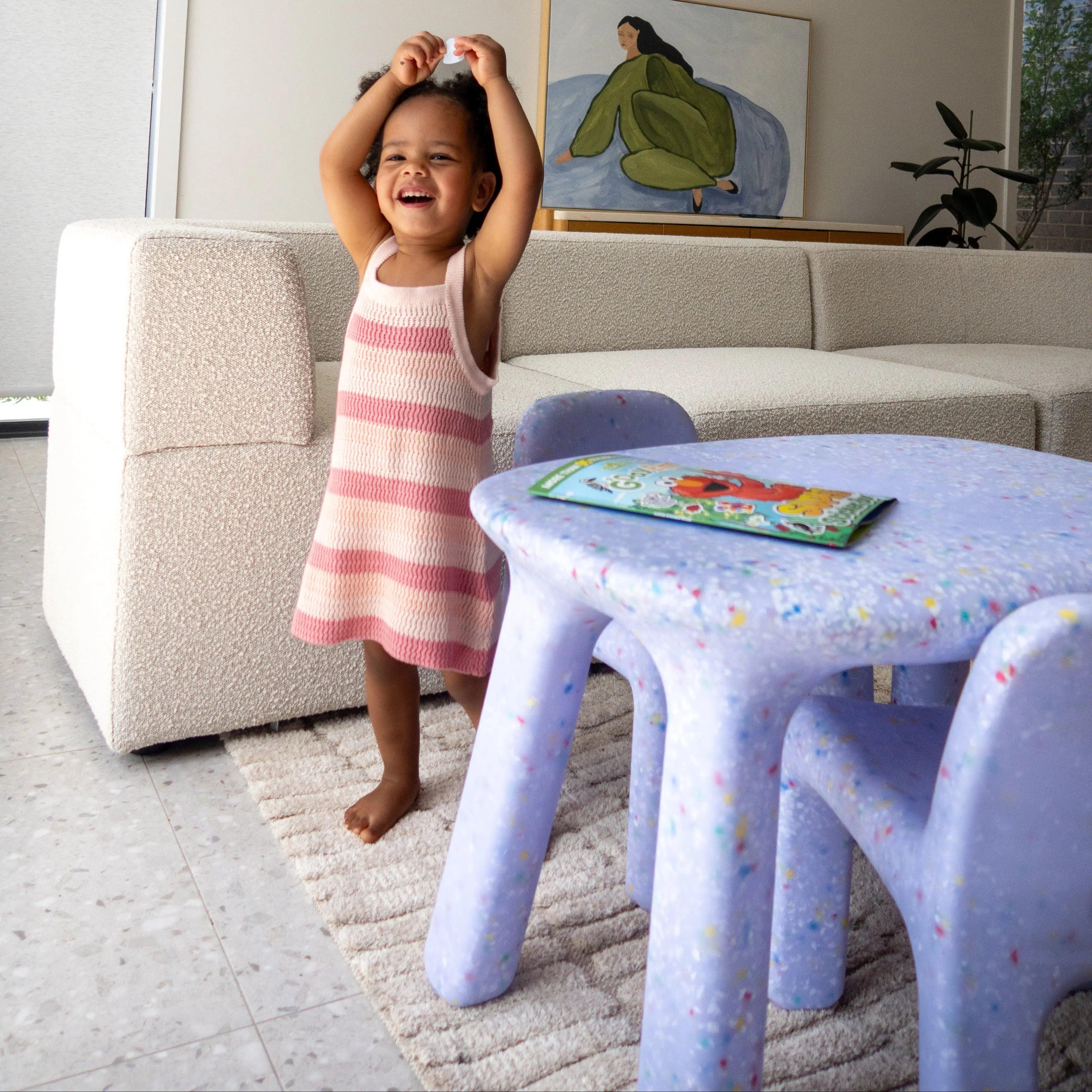 Child in a pink dress standing in a living room with a table and chairs.