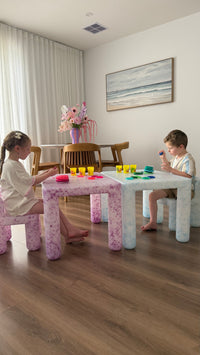 Two children sitting at a small table in a room with wooden flooring and a painting on the wall.