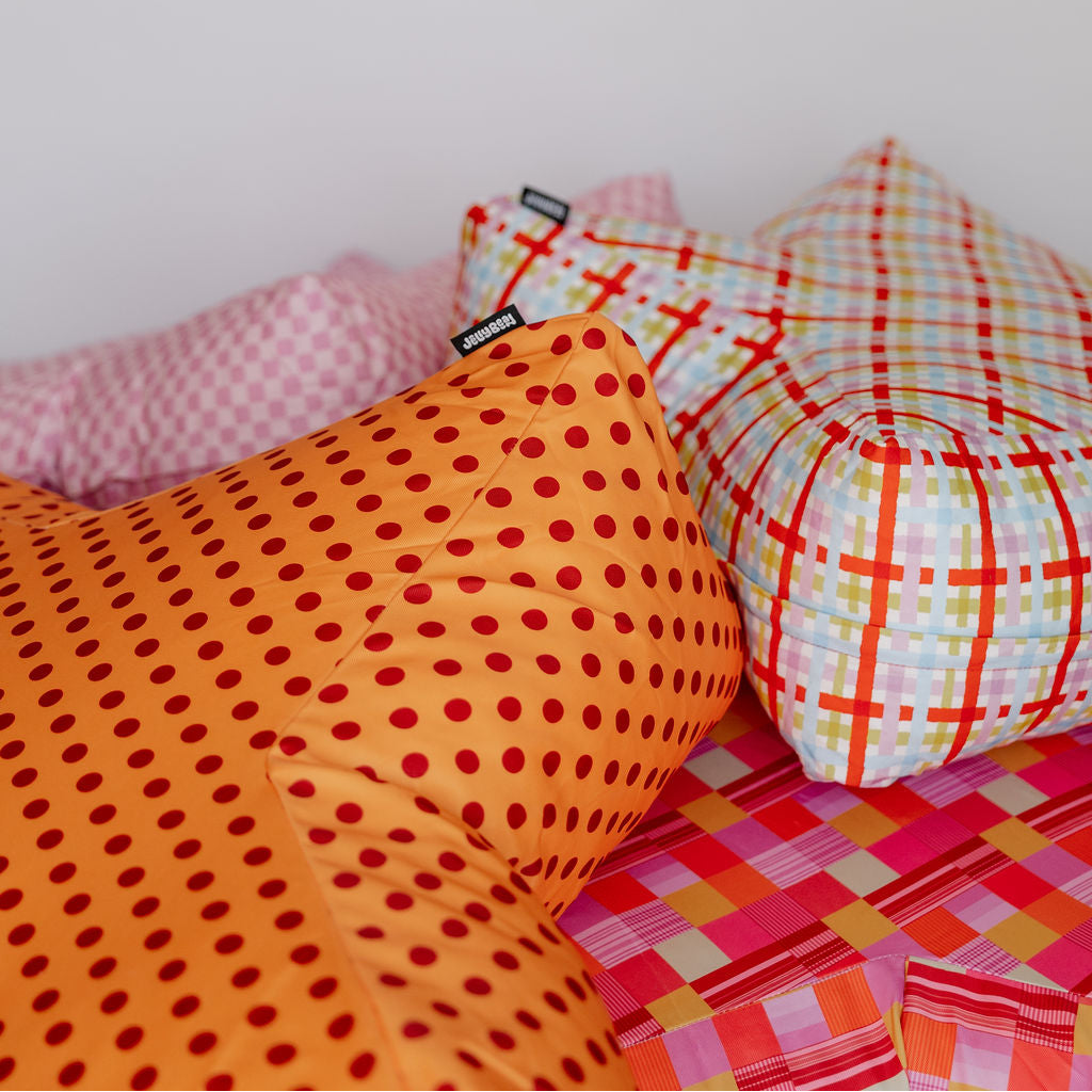 A close-up of several decorative pillows arranged on a bed against a plain light wall. The most prominent pillow in the foreground is orange with red polka dots, next to a white pillow with red and yellow plaid lines, and pink patterned pillows are visible in the background.