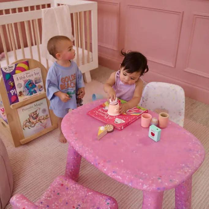 Two children playing at a pink table in a child-friendly room with toys and furniture.