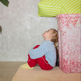 Child playing hide and seek behind a pink and white patterned box with a green striped pillow on a beige wall.