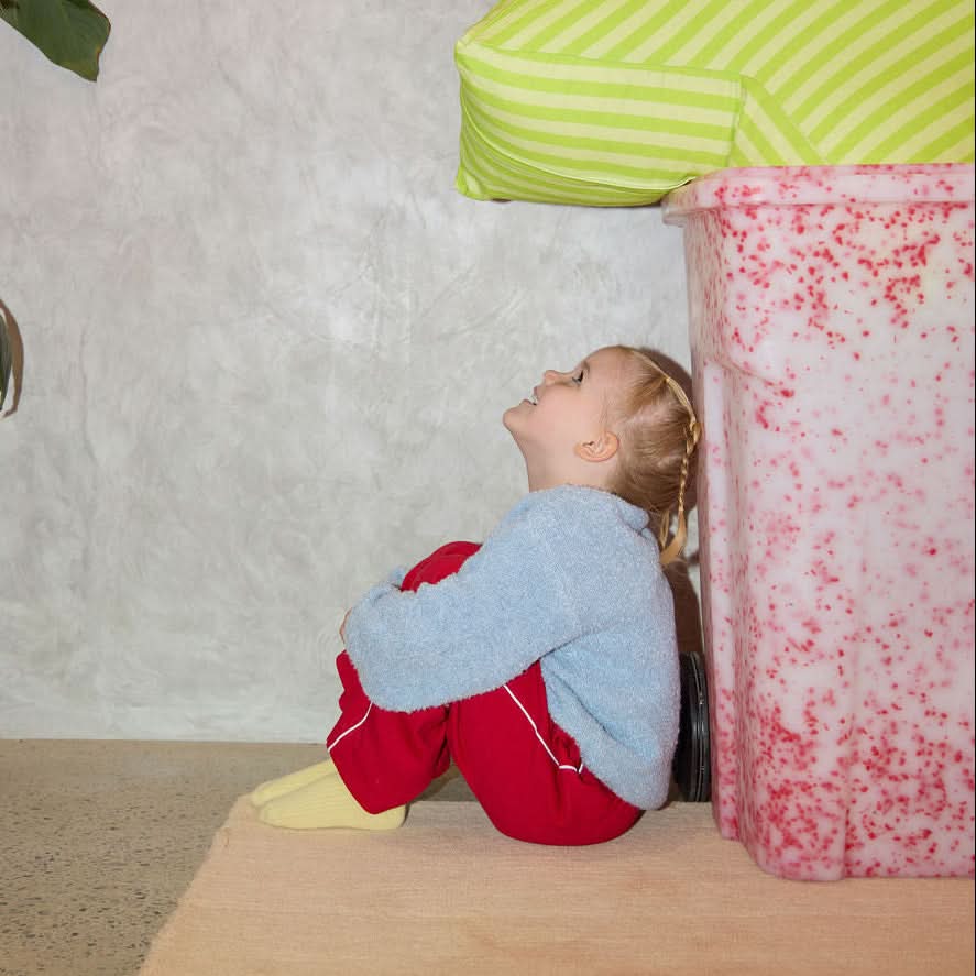 Child playing hide and seek behind a pink and white patterned box with a green striped pillow on a beige wall.