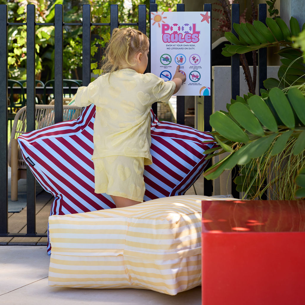 Child in yellow outfit standing on striped cushions next to a pool sign with plants and furniture in the background.