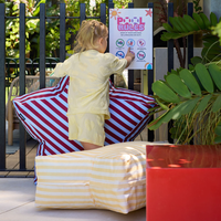 Child in yellow outfit standing on striped cushions next to a pool sign with plants and furniture in the background.